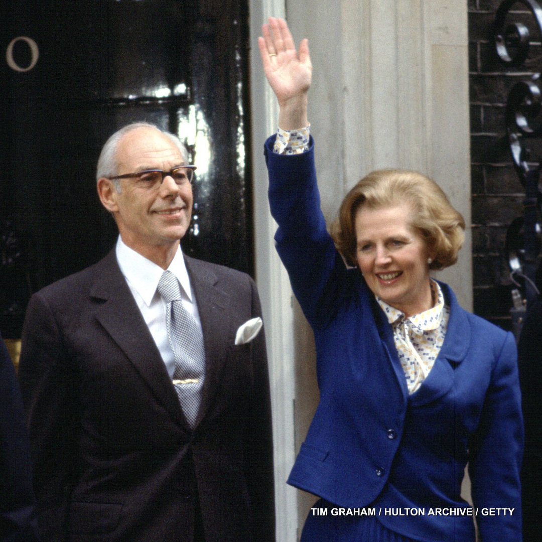 A woman in a blue suit waves and smiles beside a man in a grey suit with glasses, standing outside a black door.