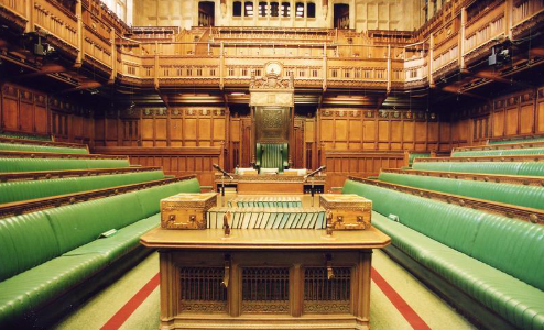 Interior of the UK House of Commons showing green benches, Speaker's chair, and despatch boxes, set in a historic, wood-panelled room.