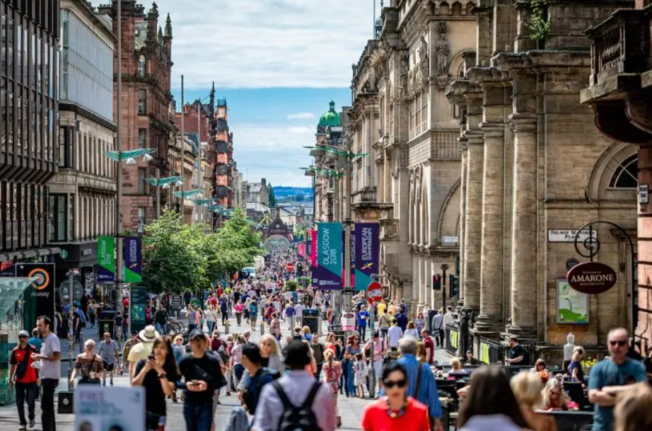 Bustling city street in Glasgow with historic architecture, colourful banners, and diverse pedestrians on a sunny day.