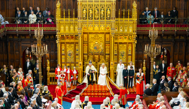 A grand ceremony in a lavish hall with golden decor and ornate thrones. Attendees in formal attire and ceremonial robes are gathered around the central figures.