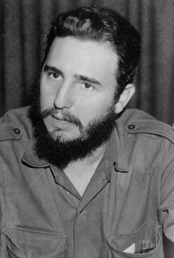 Man with a beard and military-style shirt sits against a curtained background, looking thoughtfully to the side.