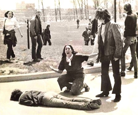A distressed woman kneels beside a prone figure on a street, surrounded by onlookers at a park, with trees and buildings in the background.