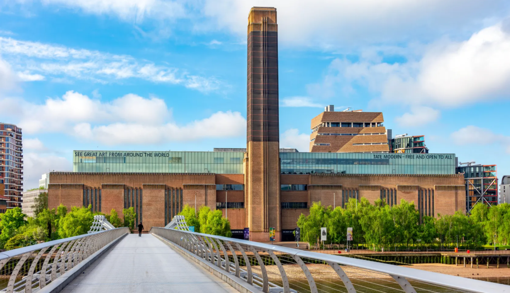 View of the Tate Modern art gallery in London, with a tall chimney, green trees in front, and a pedestrian bridge leading towards the entrance.