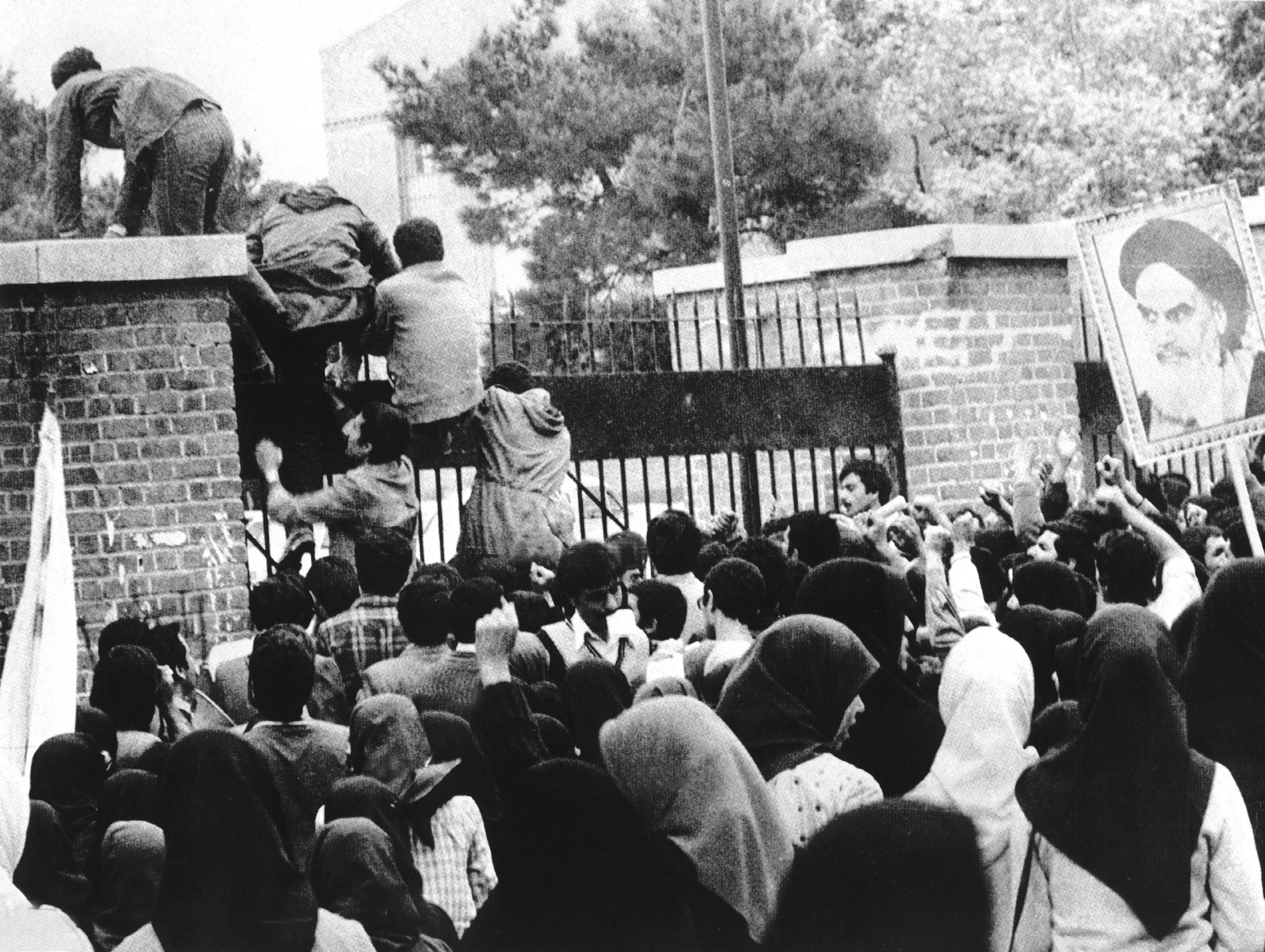 A large crowd gathers as people climb over a brick wall topped with iron fencing. A large portrait of a bearded man is held up amid the commotion.