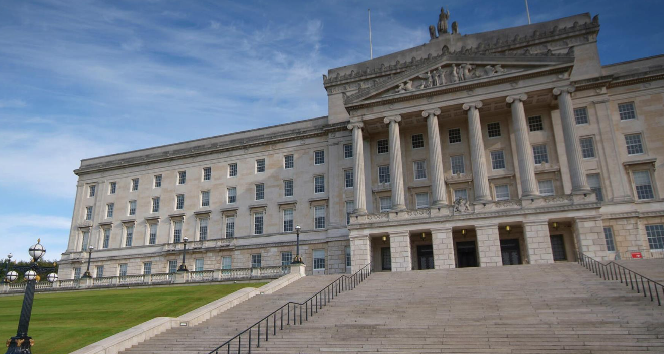 Grand neoclassical building with a wide staircase leading to its entrance, featuring tall columns and intricate sculptures, set against a blue sky.