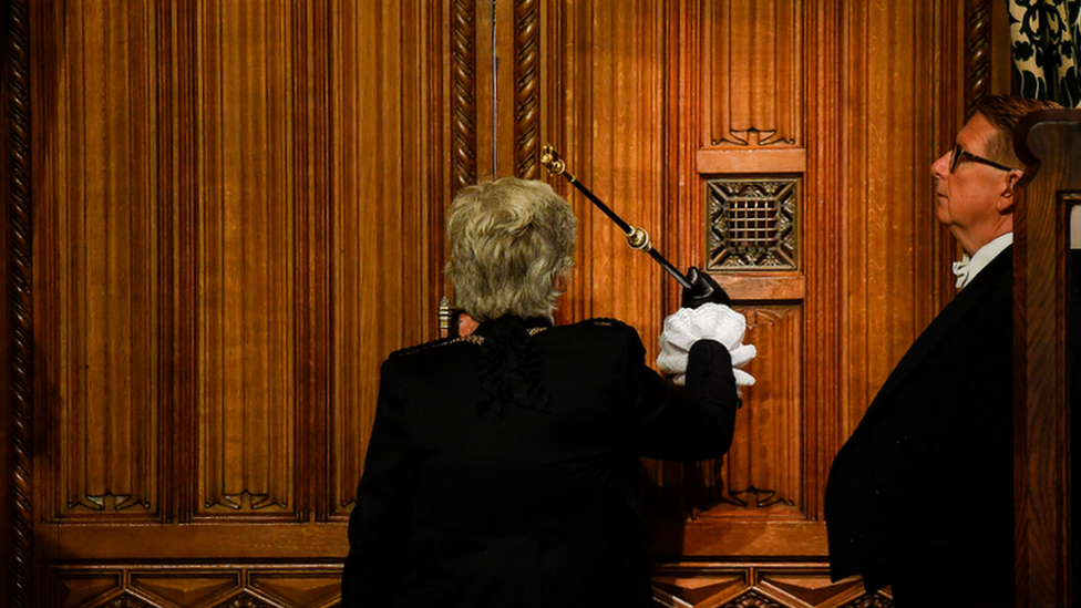 Person in formal attire holding a staff knocks on a wooden door with intricate carvings, while another person observes, in a ceremonial setting.