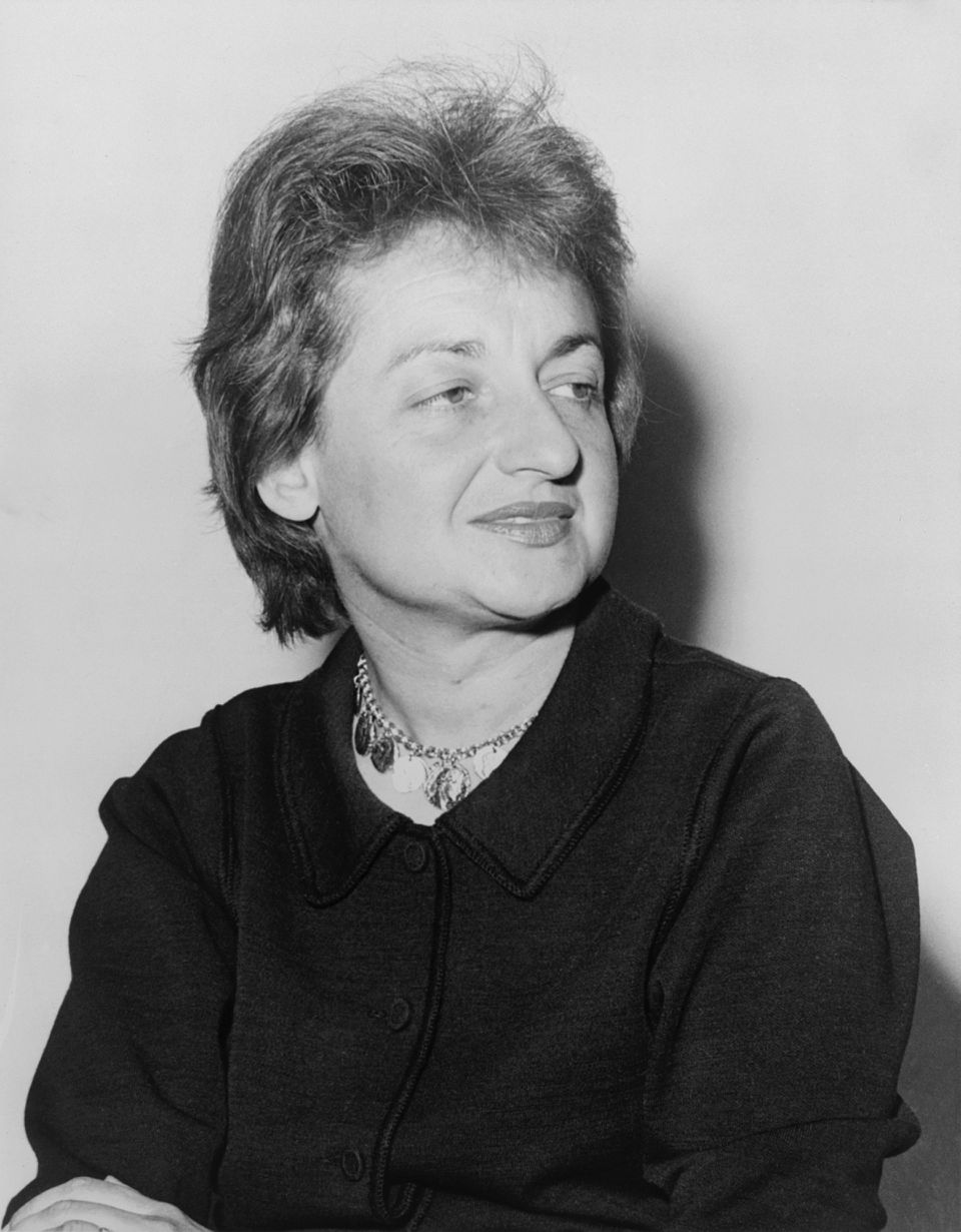 Elderly woman with short hair, wearing a dark coat and necklace, looking to the side. Plain background. Black and white portrait.