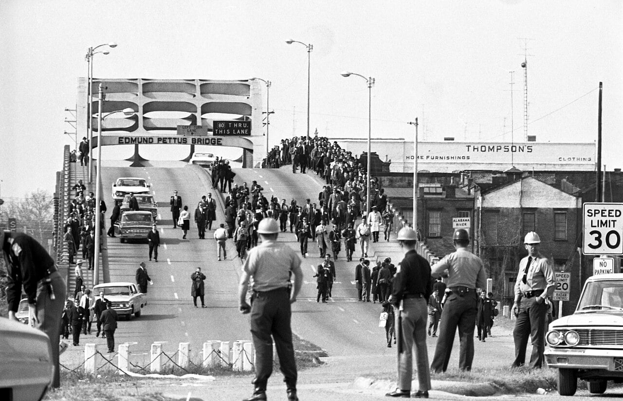A black and white image showing a large group of people walking over a bridge called the Edmund Pettus Bridge, some in suits, walking towards foreground. Policemen in helmets stand at the bottom waiting for the protestors. Buildings and vehicles visible.