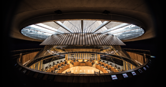 Modern circular parliamentary chamber with a large glass roof, wooden panelling, and empty seating arranged concentrically around a central podium.