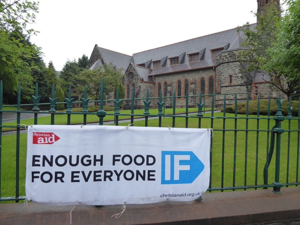 Banner on a green fence reads "Christian Aid: Enough Food for Everyone IF" in front of a stone church with lush gardens.
