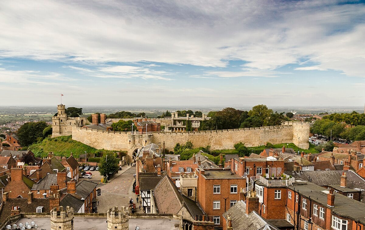 View of Lincoln Castle in England, surrounded by lush greenery and historic red-brick buildings, under a blue sky with scattered clouds.