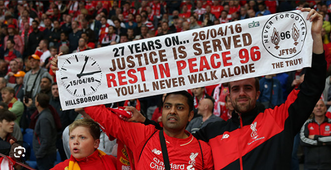 Football fans in red shirts hold a banner honouring the Hillsborough disaster victims, displaying a message of justice and remembrance.