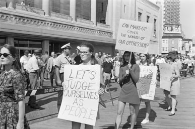 Protesters walk with signs reading "Let's judge ourselves as people" and "Can makeup cover the wounds of our oppression?" outside a large building.