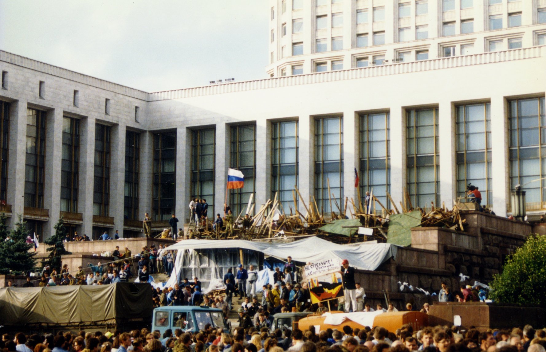 A large crowd of protesters gathers outside a large white building known as the Russian White House in Moscow during the 1991 Soviet coup attempt. People stand behind makeshift barricades of wood and tarps, with a Russian tricolor flag raised above them. Military trucks and civilians fill the foreground, while the imposing government building stands in the background.