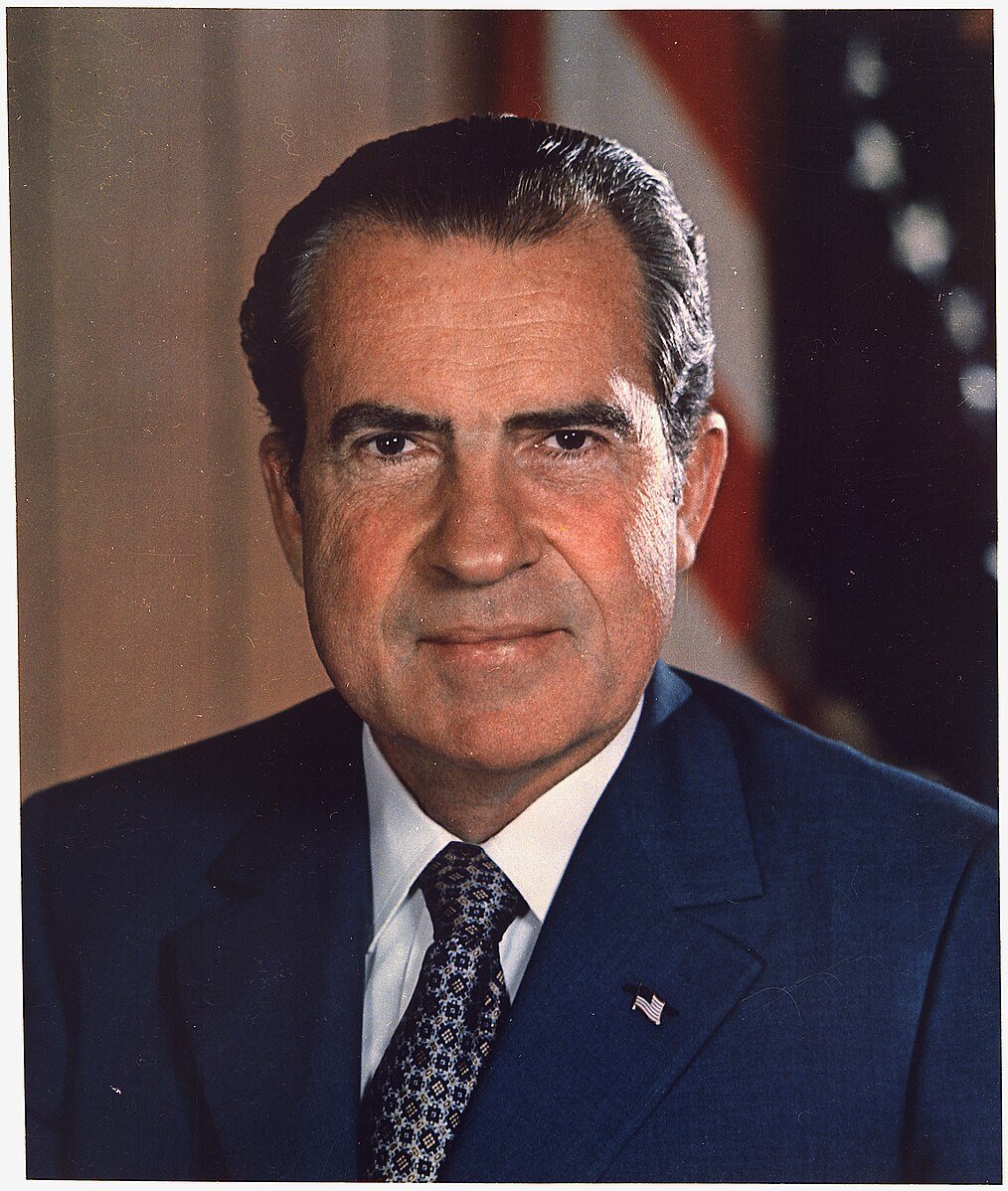 Man in a blue suit and patterned tie, with short hair, against a blurred background featuring elements of the American flag.