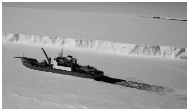 A ship navigates through icy waters near a tall, rugged ice shelf in a vast, snow-covered landscape under a clear sky.