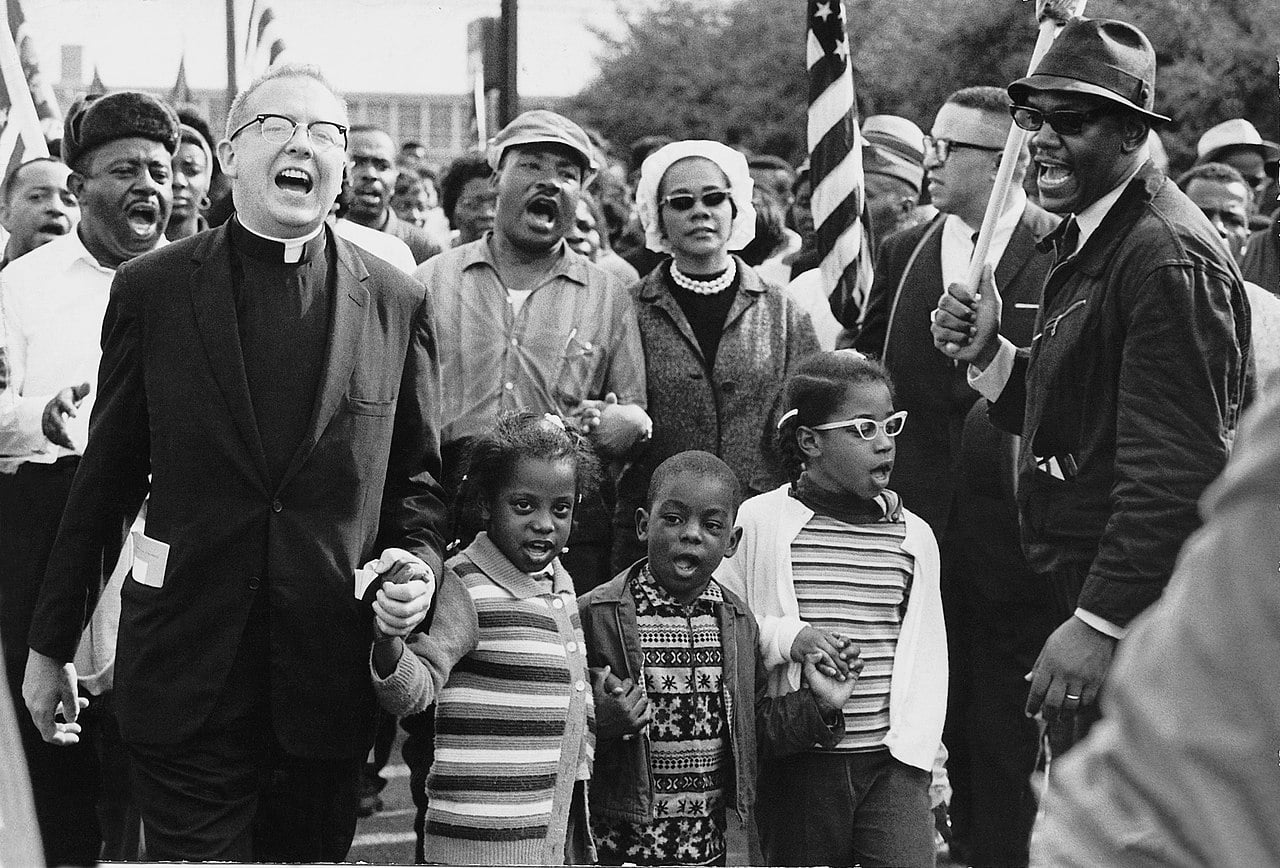 A diverse group of men, women, and children hold hands and sing during a civil rights march, with American flags visible in the background. In the middle of the photo is Martin Luther King Jr.