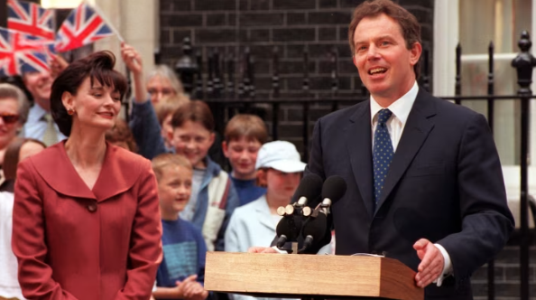 Man speaking at a podium outside a building, smiling woman beside him. Cheering crowd with Union Jack flags in the background.