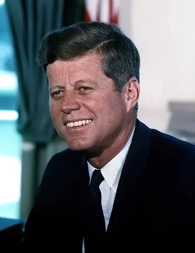 Smiling man in a dark suit and tie, seated indoors with a blurred background, featuring curtains and a partial view of a flag.