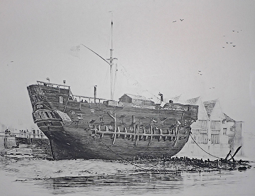 Old wooden ship docked in a harbour, surrounded by small buildings. The ship has multiple portholes and a tall mast, with calm water in the foreground.