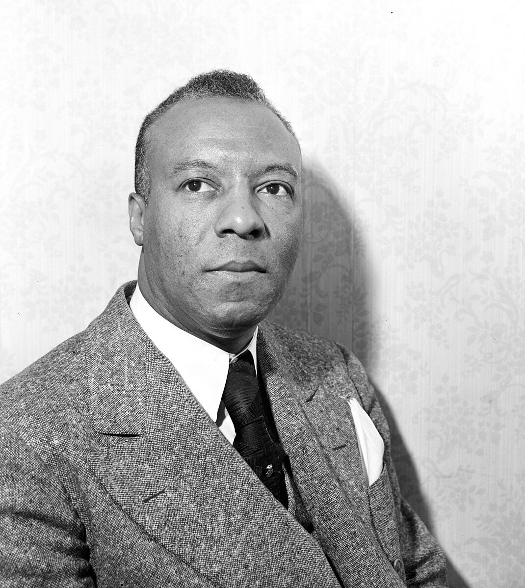 Man in a suit with a tie, looking forward against a subtle patterned background. He has a calm expression and his hair is neatly styled.
