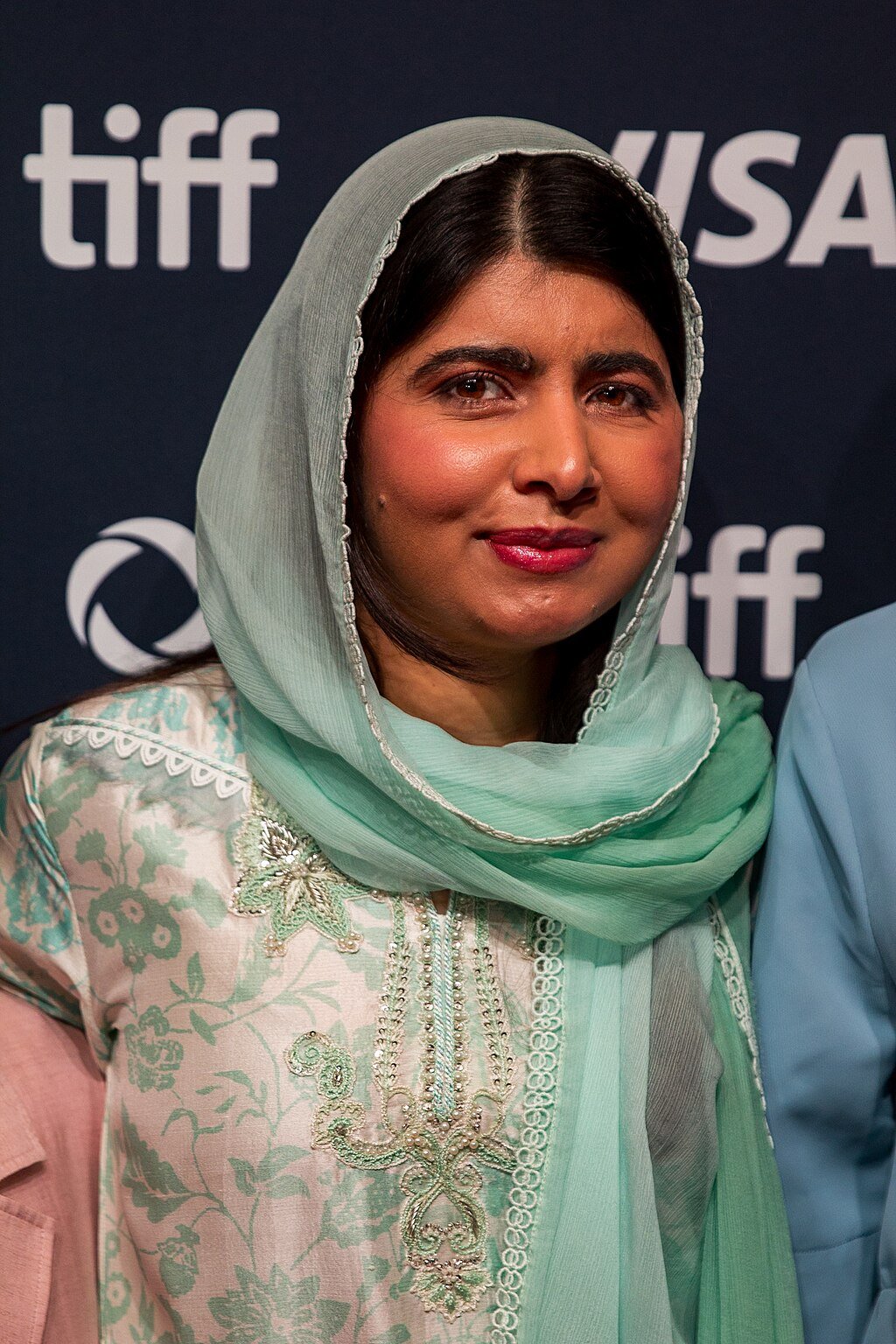 Person in a green embroidered outfit and matching headscarf, standing in front of a backdrop with logos, including "tiff" and "VISA".