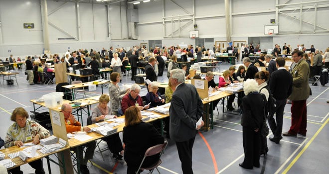 People in a large hall counting votes at numerous tables. The scene is busy with many individuals engaged in election duties under bright lighting.
