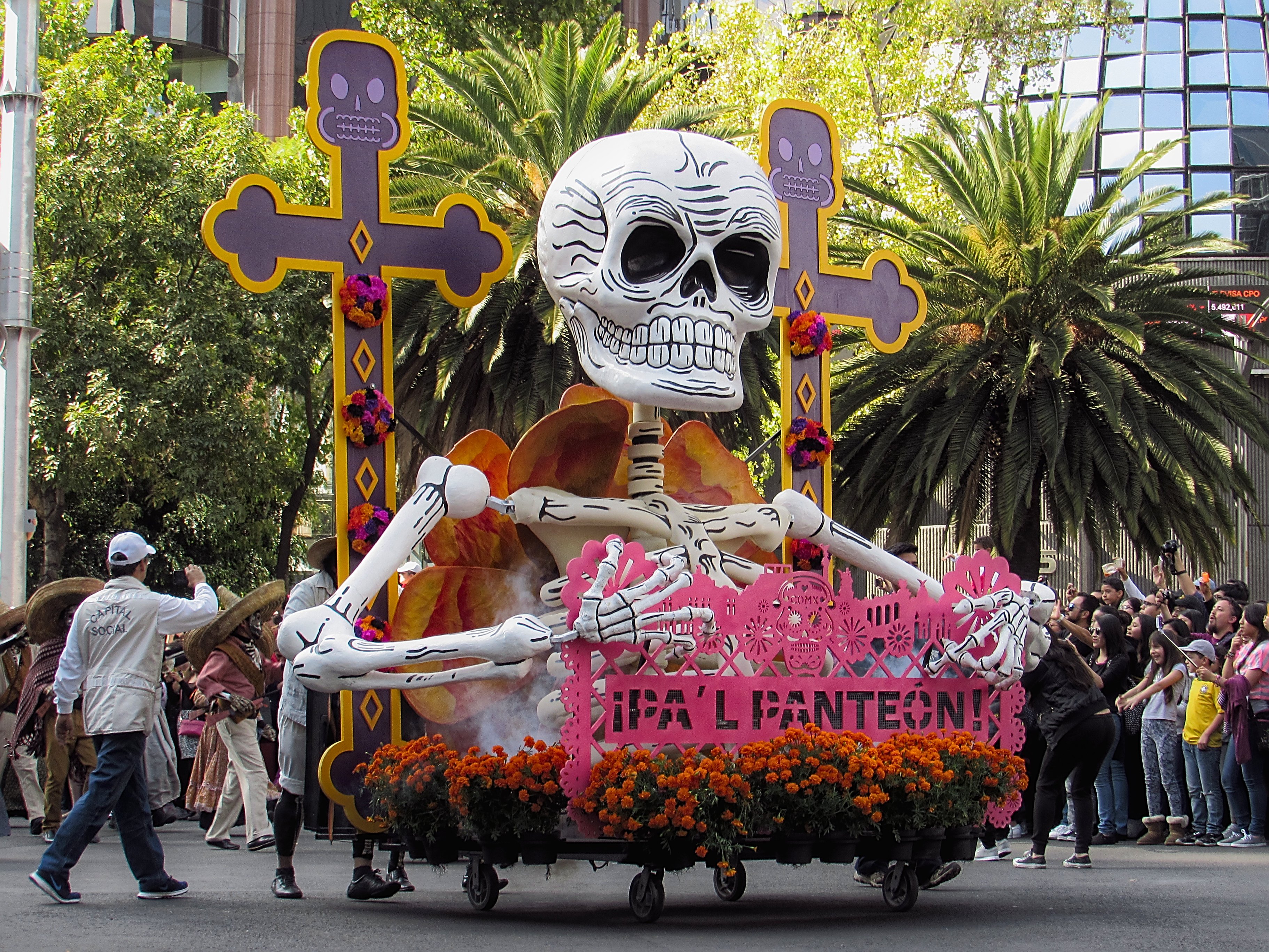 A large group of people in a town square watching colourful, giant paper mâché figures being carried in a parade