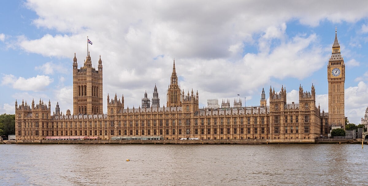 The Palace of Westminster by the River Thames, with Big Ben and Victoria Tower, under a partly cloudy sky.