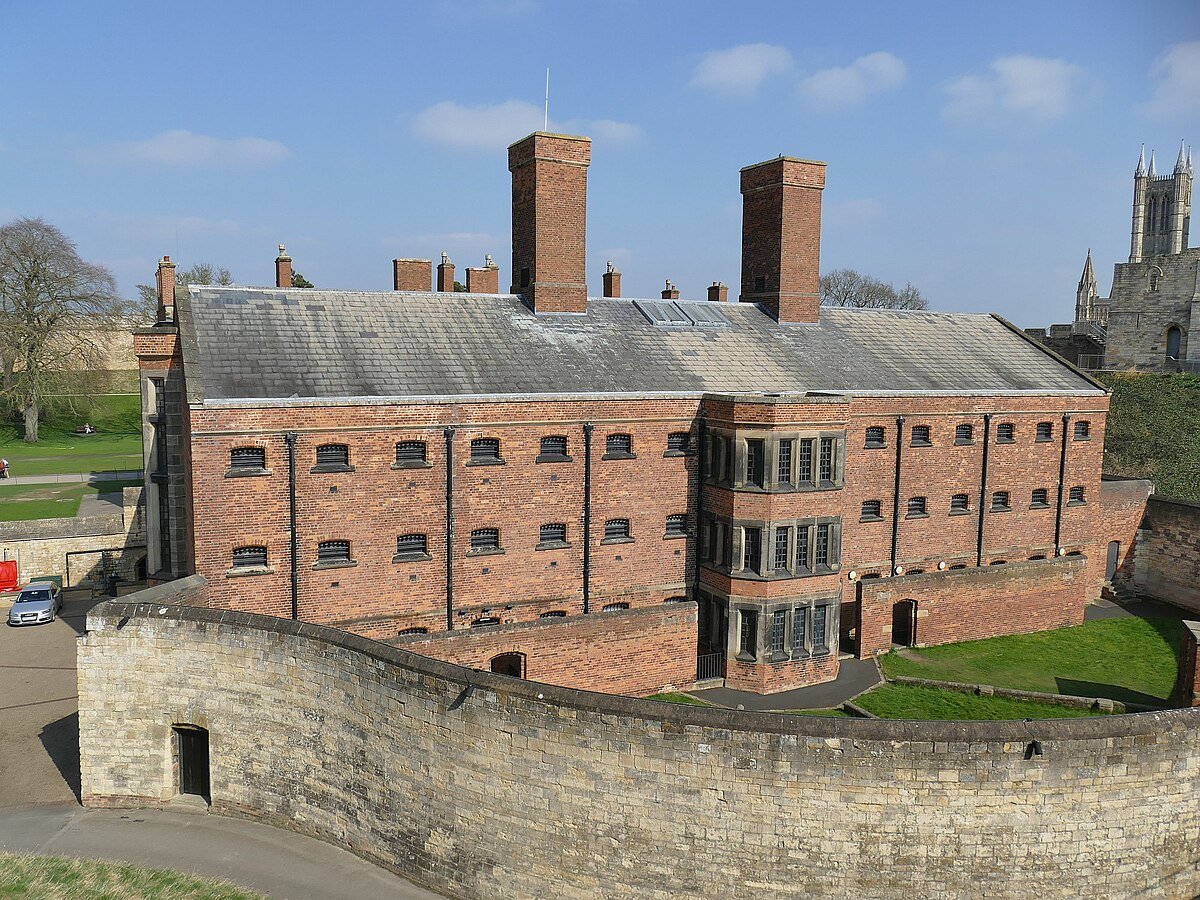 Historic red brick building with barred windows, surrounded by a high stone wall. In the background, a cathedral tower is visible under a blue sky.