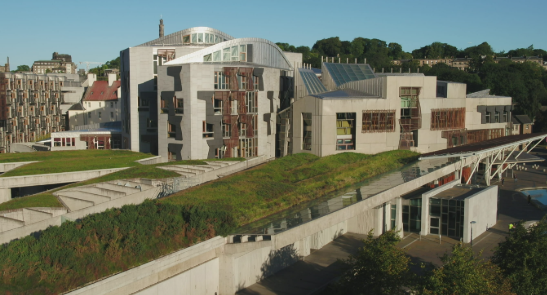 Modern building with distinct architectural design, featuring curved roofs, glass windows, and a green roof surrounded by trees on a sunny day.