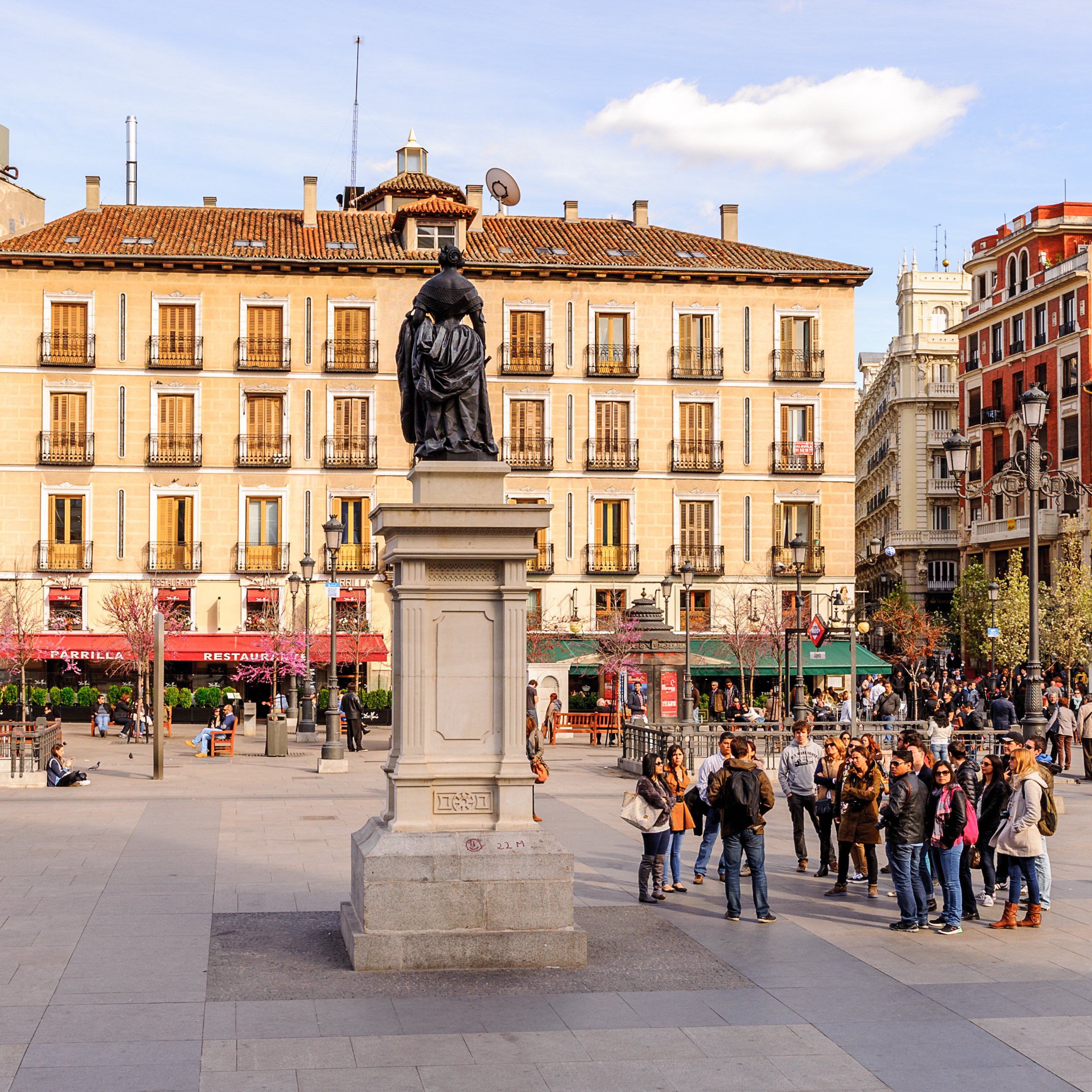 An image which shows a large, old, crowded Spanish city square with a historical statue or monument  Make sure if there’s any writing (like names of places), that it’s in Spanish. This needs to be a photo rather than an illustration.
