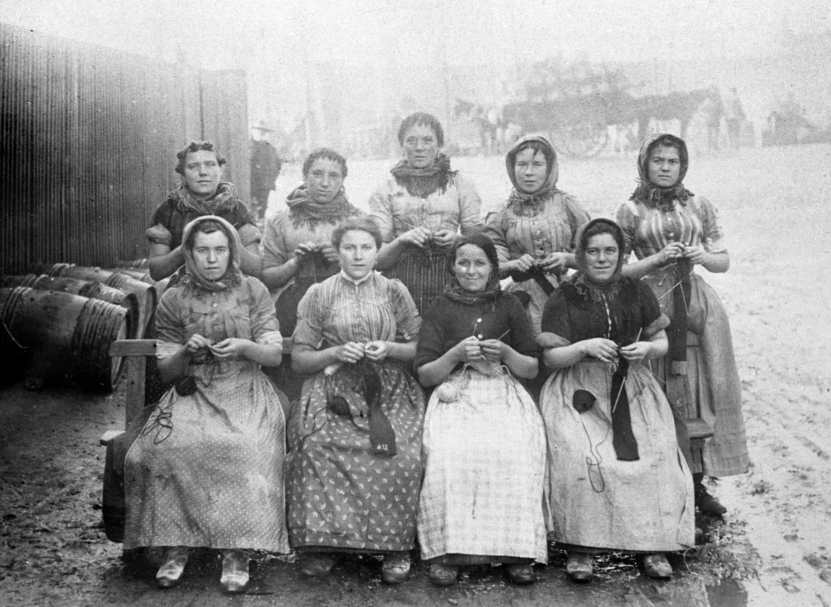 Group of nine women wearing aprons and shawls, seated outdoors; engaged in knitting; barrels and soft-focus figures in the background.