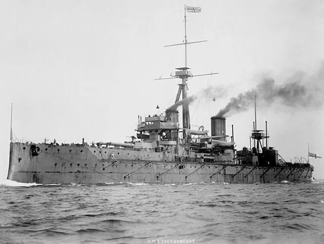 A large battleship sails on the sea, emitting smoke from its funnels. It has a tall mast and multiple gun turrets, with a flag waving atop.