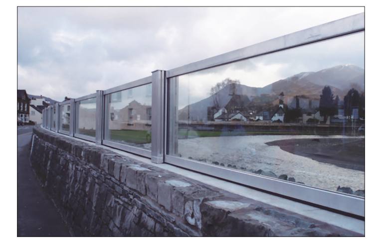 Glass barrier on a stone wall lines a riverside path with a scenic view of houses, greenery, and distant mountains under a cloudy sky.