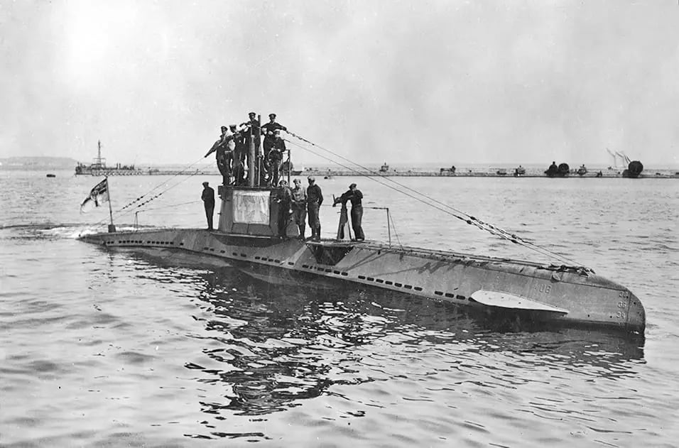 Historic submarine with several crew members standing on the deck in a harbour, flying a naval flag, with a coastline and dock structures in the background.