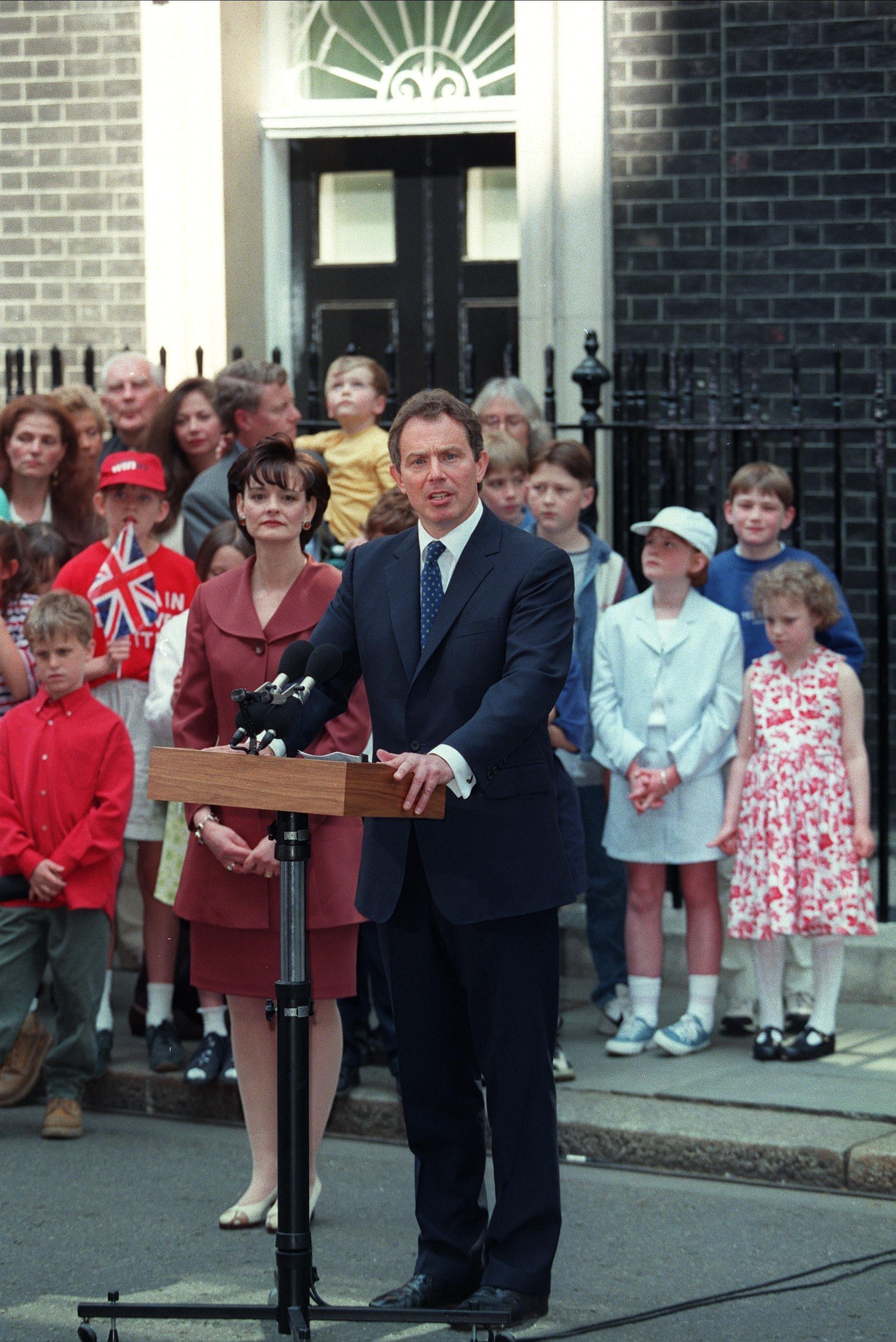 A man in a suit speaks at a podium outside a building, surrounded by a diverse crowd including children and adults, one holding a Union Jack flag.