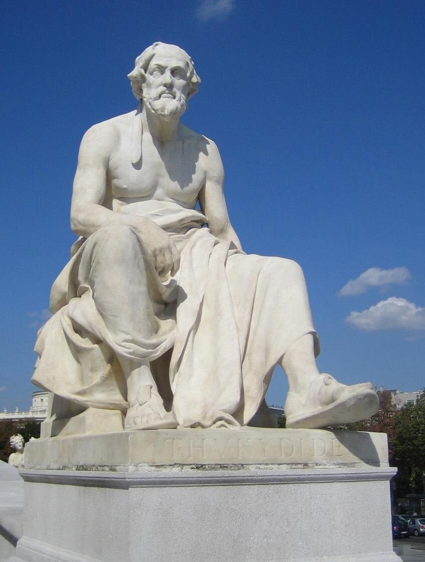 Marble statue of a seated, bearded man in a toga against a clear blue sky, with trees and buildings in the background.