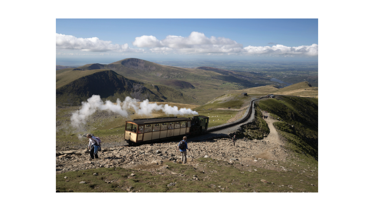 Steam train ascending a mountain with hikers on a rocky path. Scenic view of vast landscape under a blue sky with scattered clouds.