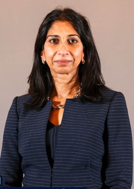 Woman with long dark hair wearing a navy blazer, standing against a plain light brown background, looking directly at the camera.