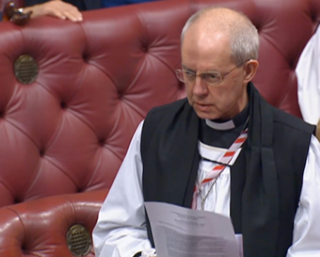 Bald man in clerical attire reads a document while seated on a red, tufted bench with brass buttons in a formal setting.