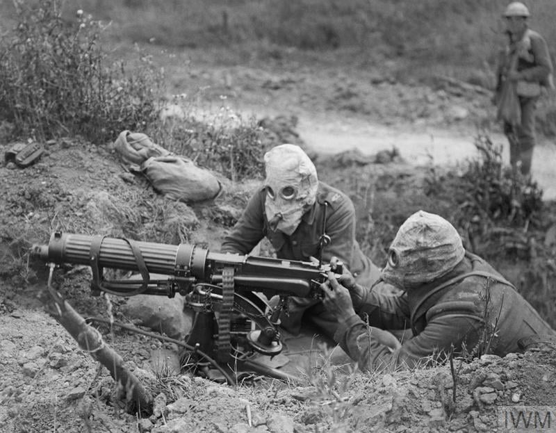 Soldiers wearing gas masks operate a machine gun in a trench during wartime, with one man standing in the background, surrounded by vegetation.