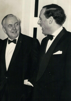 Two men in formal black-tie attire stand indoors, engaging in conversation. One is smiling warmly; both wear dark suits with bow ties and white shirts.