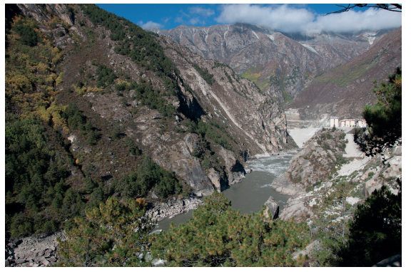 Mountainous landscape with a winding river cutting through steep, rocky slopes, lined with scattered trees under a blue sky with few clouds.