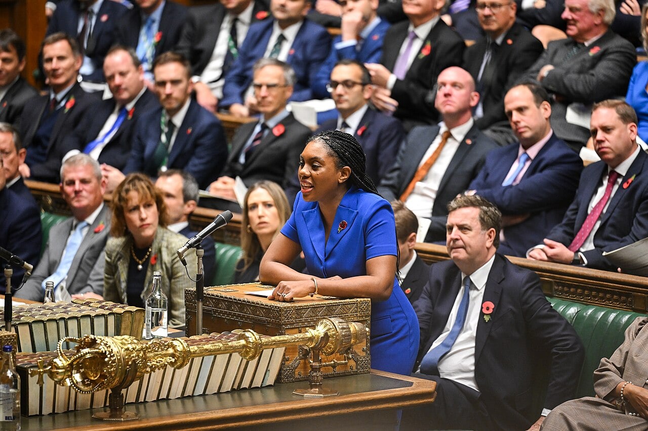 A woman in a blue dress speaks at the UK House of Commons, with seated members wearing poppies, denoting a formal parliamentary session.