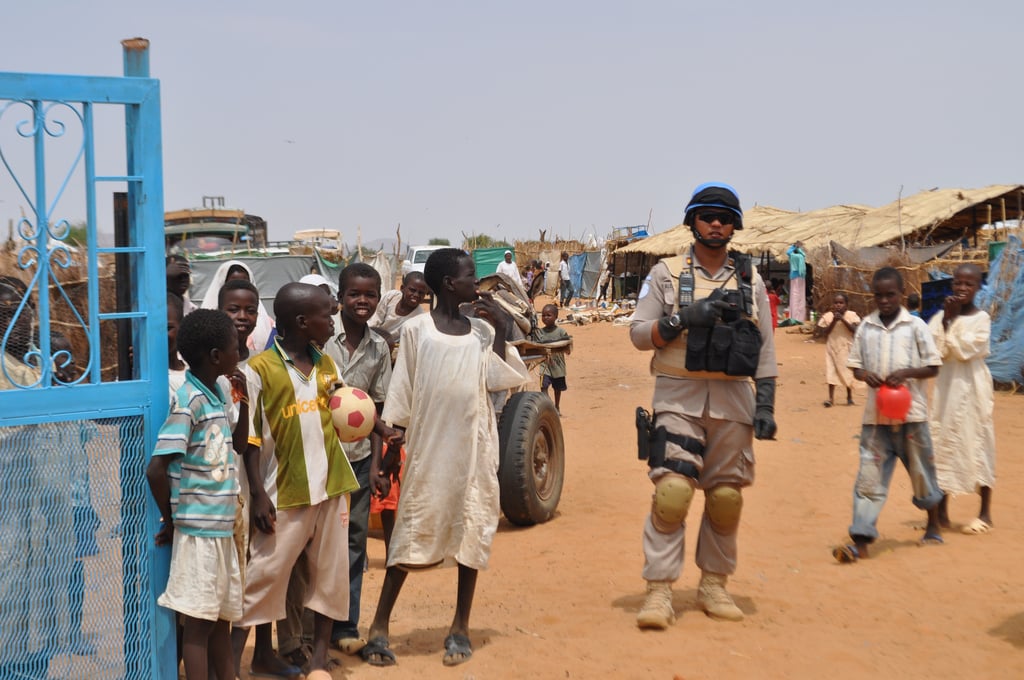A peacekeeper in uniform patrols a rural area with children nearby; one holds a football. Tents and huts are visible in the background.