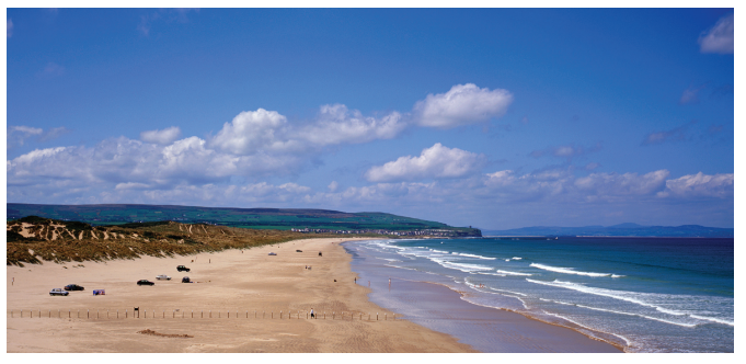Sandy beach stretching along a coastline with gentle waves, sparse clouds in a blue sky, and several parked cars on the sand near grassy dunes.