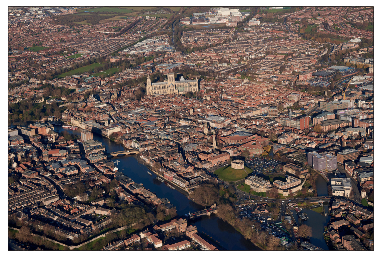 Aerial view of a historic city featuring a large cathedral at its centre, surrounded by densely packed buildings and a river with several bridges.