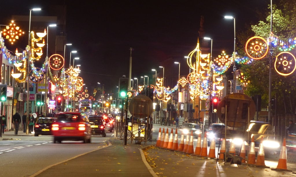 Busy street at night adorned with festive lights, trailing car lights, traffic signals, and orange cones line the road; pedestrians on the pavement.