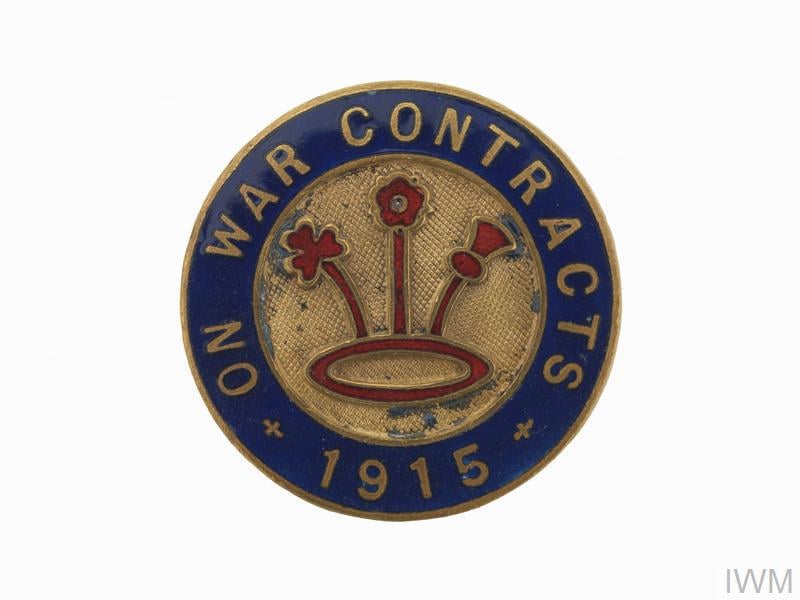 Circular 1915 badge with the words "On War Contracts" on a blue border and a central design of three red flowers on a gold background.